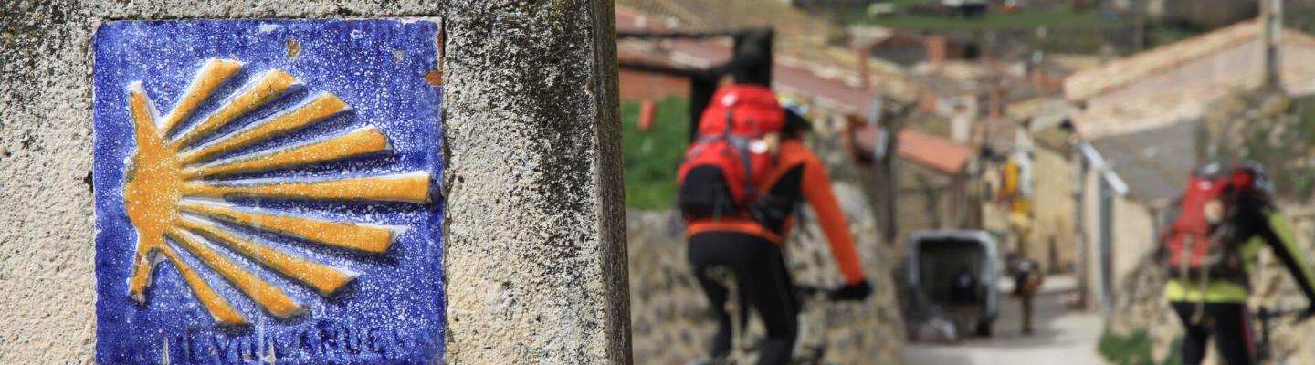 Muschel-Wegweiser auf dem Jakobsradweg mit zwei Radreisenden | © adobe.stock - (c)alecki